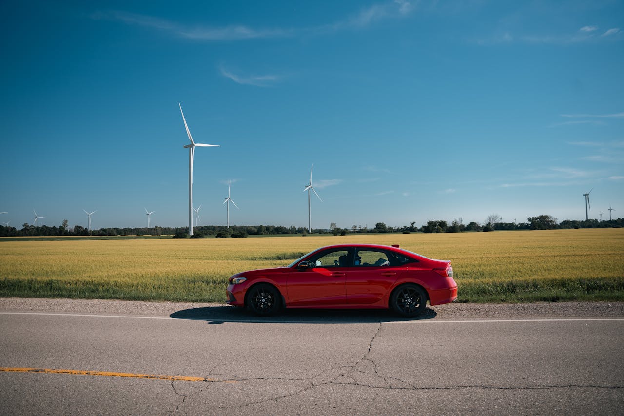 A red sedan on a rural road beside fields with wind turbines under a clear blue sky.