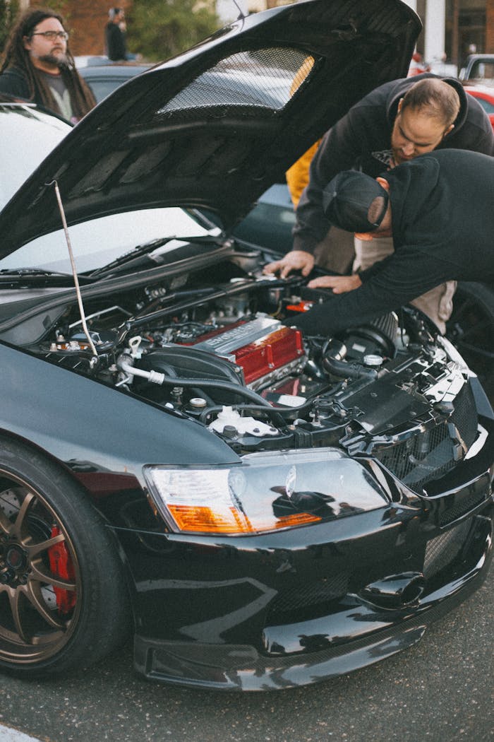Men examining the engine of a black car with hood open at an outdoor auto show.
